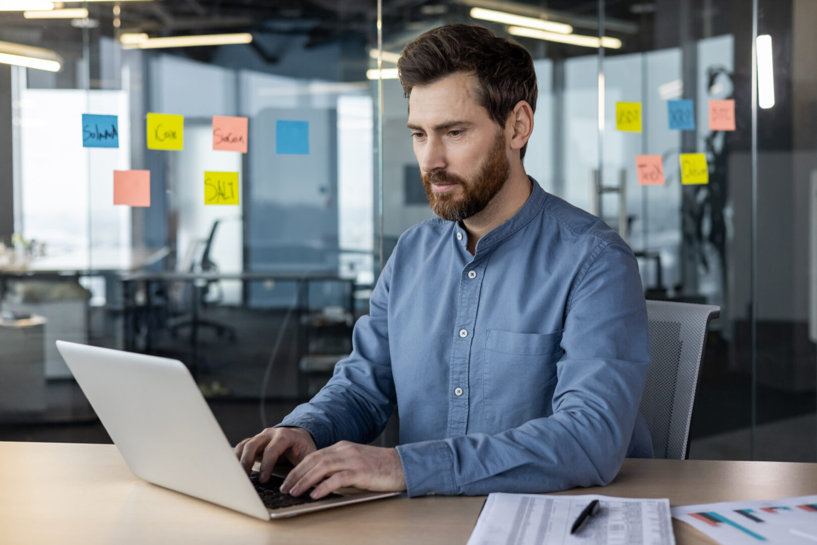 Focused businessman working on laptop in modern office with glass partitions and sticky notes