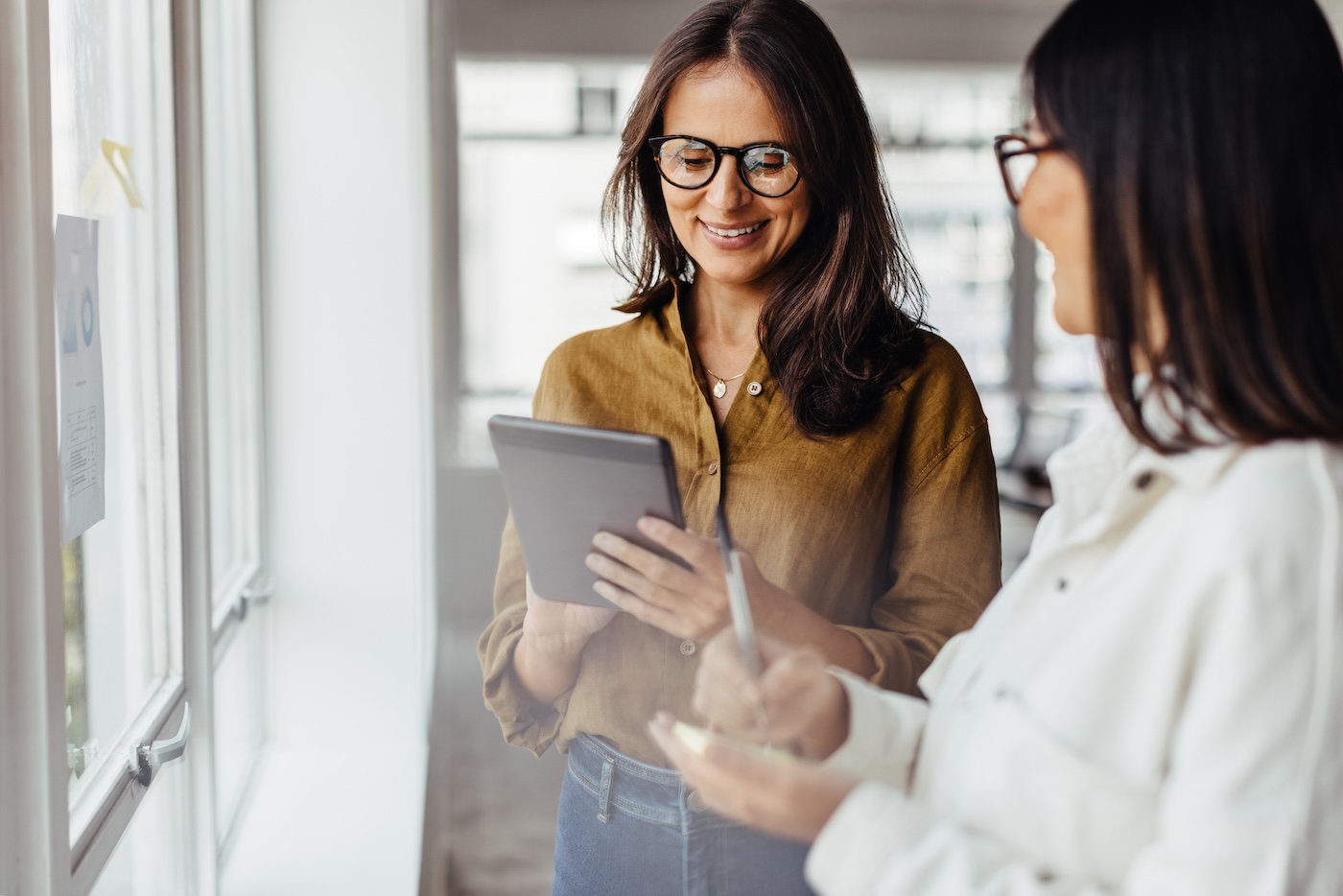 Business women standing next to a window and discussing their ideas. Female business professionals brainstorming using stocky notes and a tablet.